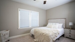Ein Schlafzimmer mit einem Bett, einem Nachttisch, einer Lampe, einem Deckenventilator und einem Fenster mit Jalousien.