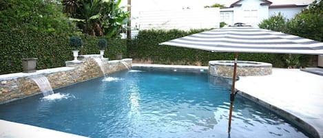 A swimming pool with a stone border, a waterfall feature, and a striped umbrella.
