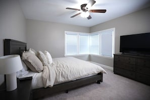 Ein Schlafzimmer mit Bett, Deckenventilator, Kommode, Fernseher und Fenster mit Jalousien.