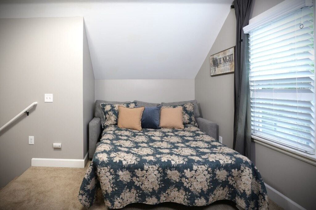 Un dormitorio con cama, ventana con persianas, interruptor de luz en la pared y un cuadro enmarcado.