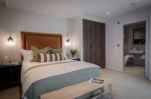 A modern bedroom with a large bed, bedside lamps, a nightstand, a bedside table, a wooden bench, and a view into a bathroom.