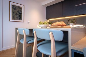 A modern kitchen with dark cabinets, a light countertop, and a bowl of bananas. There are two blue chairs and a small plant on the counter.