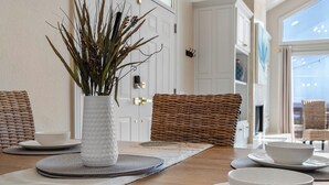 Une table à manger avec des bols blancs, un vase blanc contenant des plantes séchées et des chaises en osier.