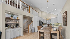 Une cuisine spacieuse avec des meubles blancs, un coin repas doté de chaises en osier et un escalier menant à l’étage supérieur.