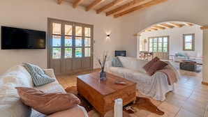 A spacious living area with a wooden coffee table, two white sofas, a TV mounted on the wall, and a large glass door leading to an outdoor area.