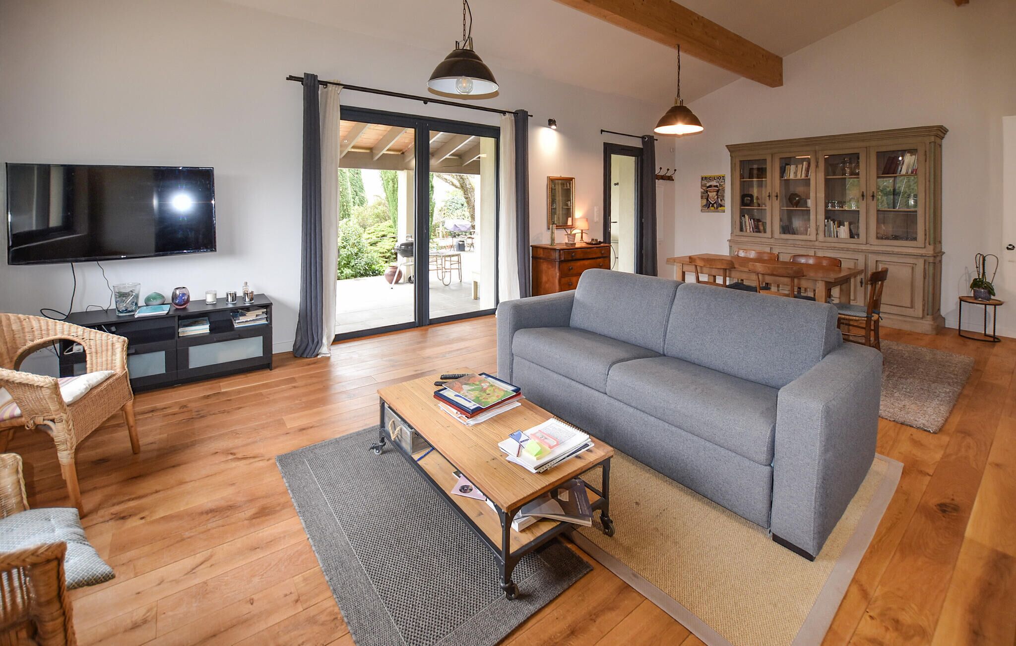 A living room with a grey sofa, wooden flooring, a TV on the wall, a dining area with a table and chairs, and a cabinet with glass doors.