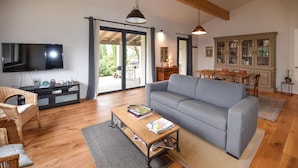 Un salon avec un canapé gris, un plancher en bois, un téléviseur fixé au mur, un coin repas avec une table et des chaises, et un meuble avec des portes en verre.