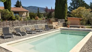 Une piscine rectangulaire bordée de pierres, entourée d’un muret en pierre et d’une clôture en bois. Plusieurs chaises longues sont alignées le long du muret.