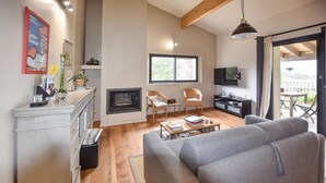 Un salon avec une cheminée, deux fauteuils en osier, un canapé gris, une table basse et un téléviseur fixé au mur.