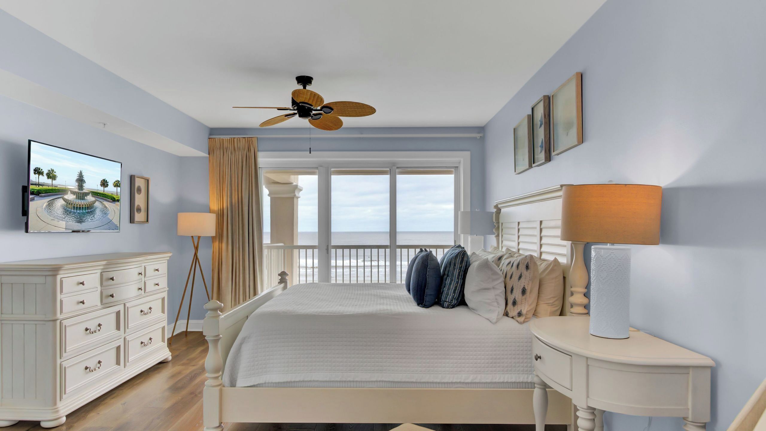 A bedroom with a bed, bedside tables, a ceiling fan, a large window with a view, and a wall-mounted TV.