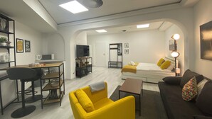 Una sala de estar moderna con un sillón amarillo, un sofá marrón y una cama con almohadones amarillos. Hay un escritorio con una silla y un televisor sobre un mueble bajo. La habitación tiene paredes blancas y piso de madera.