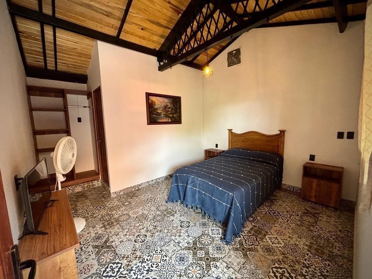 Een slaapkamer met een bed, een houten hoofdbord, een ventilator, een bureau en vloertegels met een patroon.