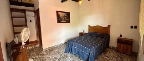 Een slaapkamer met een bed, een houten hoofdbord, een ventilator, een bureau en vloertegels met een patroon.