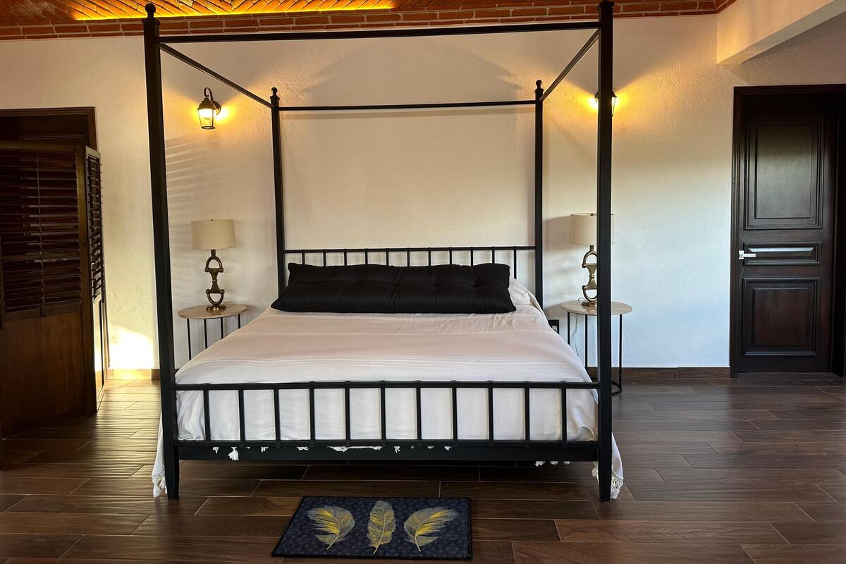 A four-poster bed with a white mattress and black headboard, flanked by two bedside tables with lamps, in a room with wooden flooring and a brick ceiling.