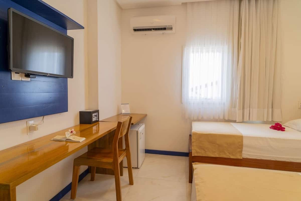Une chambre d’hôtel avec un bureau en bois, un téléviseur fixé au mur, un petit climatiseur, un lit recouvert d’une courtepointe blanche et une fenêtre avec des rideaux légers.