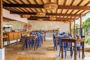 Un área de comedor al aire libre con mesas de madera y sillas azules, un muro de piedra al fondo y lámparas de mimbre colgantes.