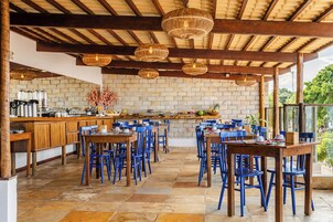 Un área de comedor al aire libre con mesas de madera y sillas azules, un muro de piedra al fondo y lámparas de mimbre colgantes.
