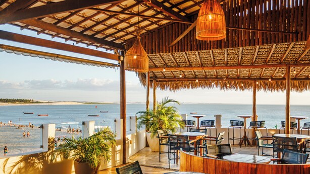 Un patio frente a la playa con mobiliario exterior, vista al mar y una estructura de techo de paja.