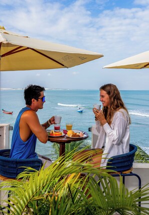 Two people sitting at a table with a view of the ocean, a boat in the distance, and a tropical setting with plants. - VILA GALLINA POUSADA (Jijoca de Jericoacoara)
