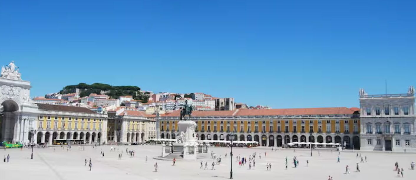 Una amplia plaza abierta con edificios históricos, una fuente y una colina con árboles al fondo.