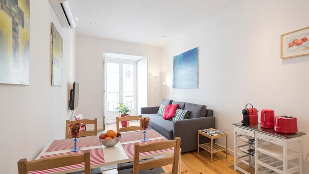 A modern living room with a grey sofa, a dining table with chairs, a coffee table, and a kitchen area with a white cabinet and red appliances.