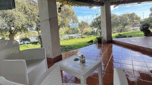 An outdoor patio with white wicker furniture, a tiled floor, and a view of a green landscape with trees and a clear sky. - DOMO MEA beautiful farmhouse immersed in theSardinian countryside an its animals (Nulvi)