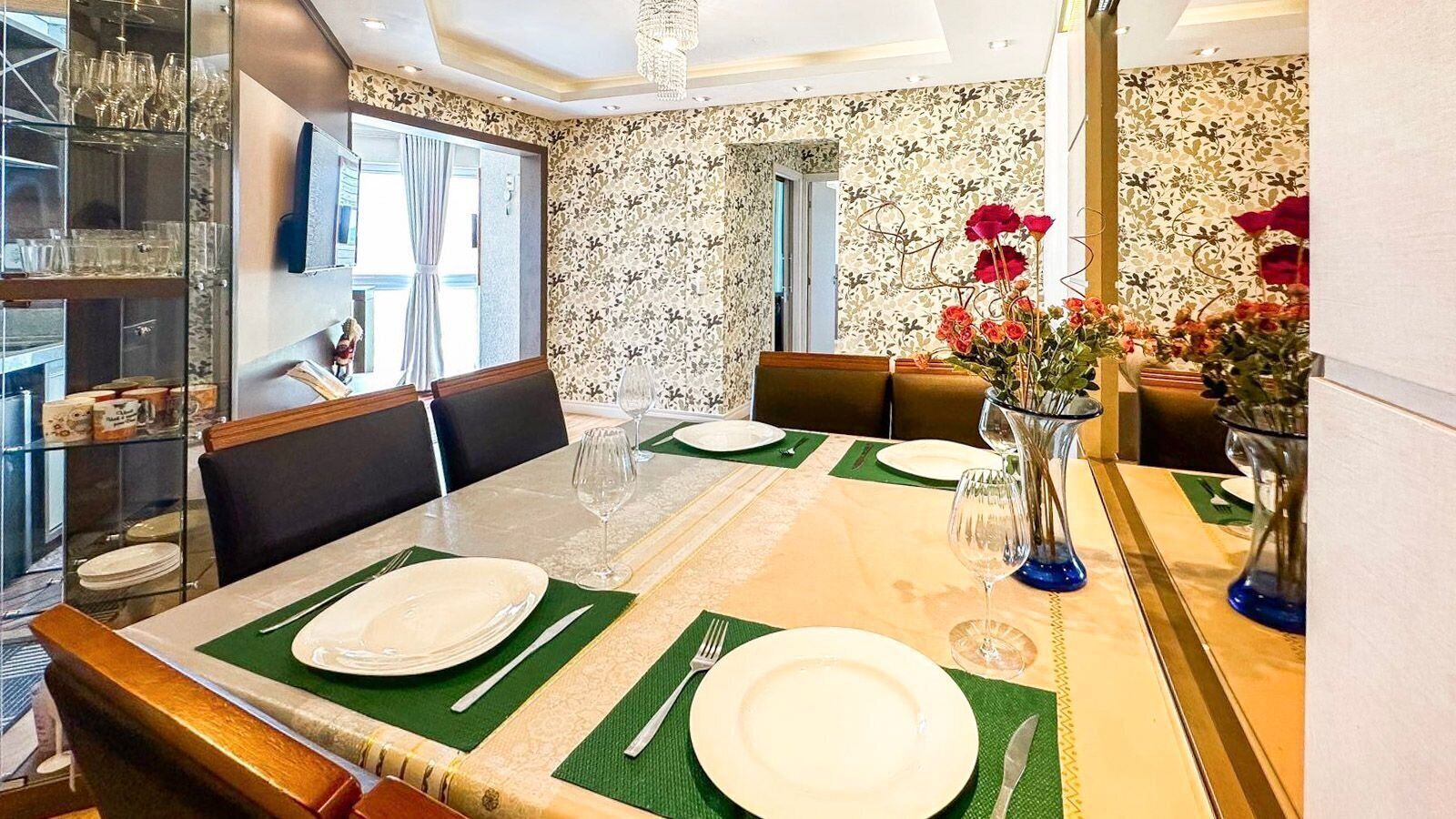 Un comedor con una mesa preparada para cuatro, sillas, un florero con flores y un aparador con copas de vino.