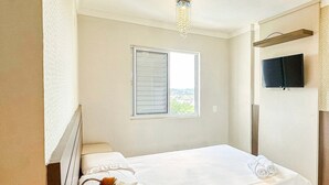 A single bed with white bedding, a bedside table with a lamp, a window with blinds, and a television on the wall. - Resid. Clube Mar Grosso-Laguna - LAG905 (Laguna)