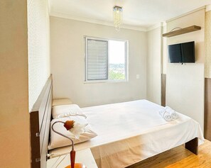 A single bed with white bedding, a bedside table with a lamp, a window with blinds, and a television on the wall. - Resid. Clube Mar Grosso-Laguna - LAG905 (Laguna)