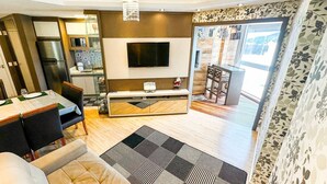 A modern living room with a sofa, a dining area with a table set for two, a kitchen area with a refrigerator and a TV mounted on the wall, and a large window providing natural light. - Resid. Clube Mar Grosso-Laguna - LAG905 (Laguna)