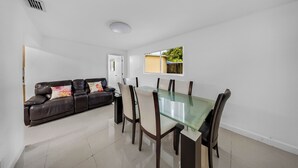 A modern living room with a glass dining table, leather sofa, and a view of a garden through a window.