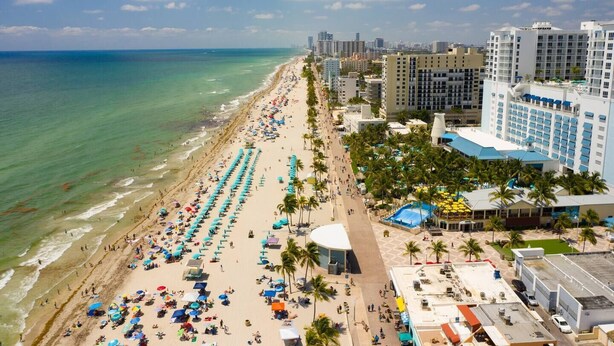 A beachside hotel with a long sandy beach, a pool area, and several high-rise buildings in the background.
