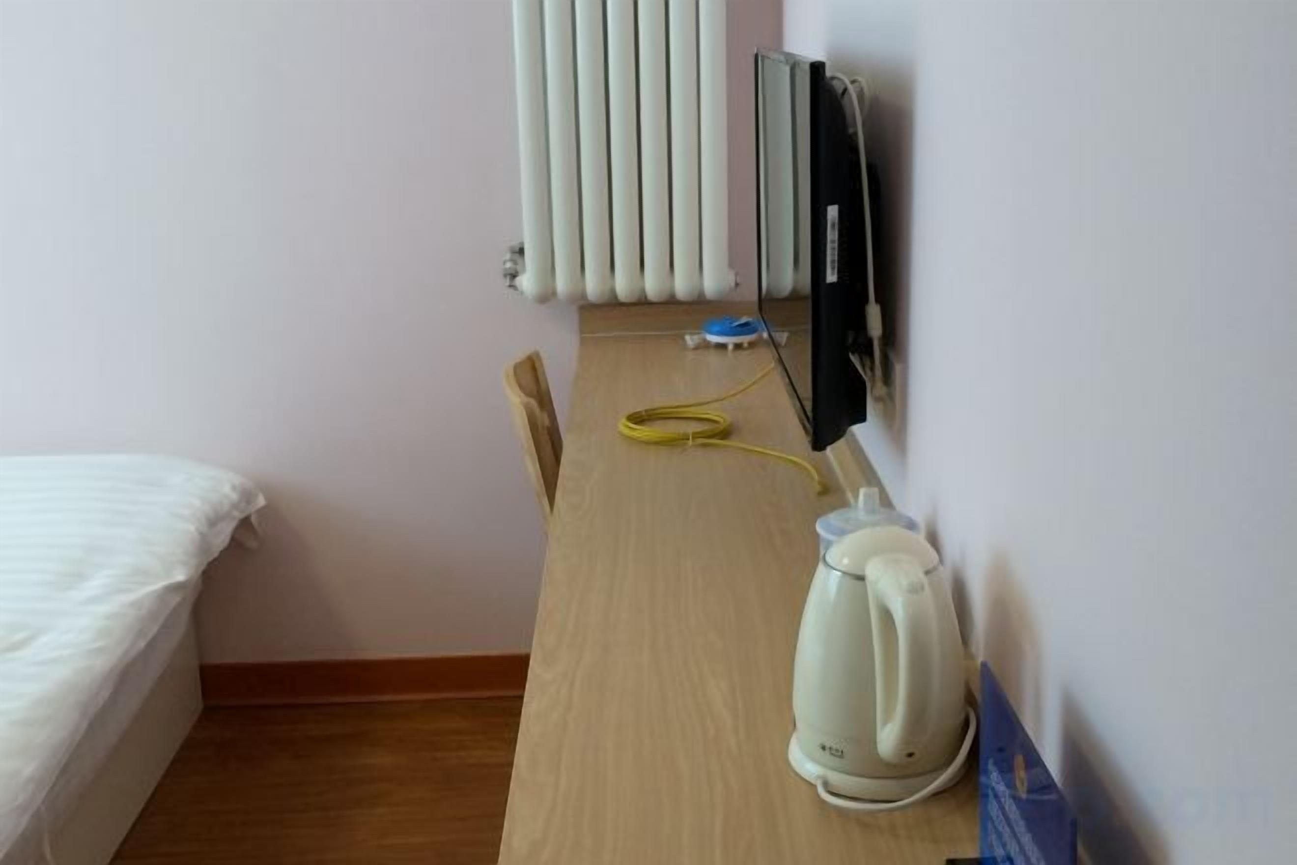 A hotel room with a bed, a radiator, a kettle, and a desk with a chair.