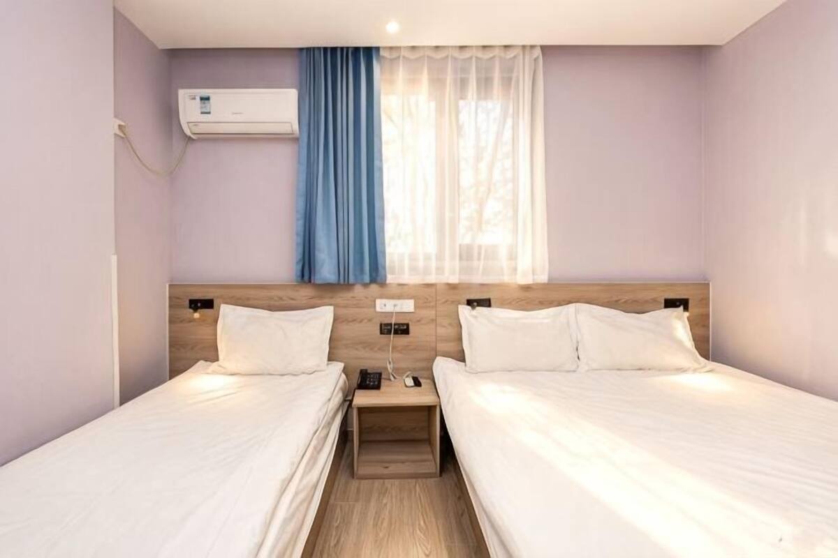 A twin room with two beds, a wooden headboard, a nightstand, an air conditioning unit, and a window with sheer curtains.