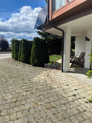 A paved outdoor area with a solar panel, a seating area with chairs, and neatly trimmed hedges.