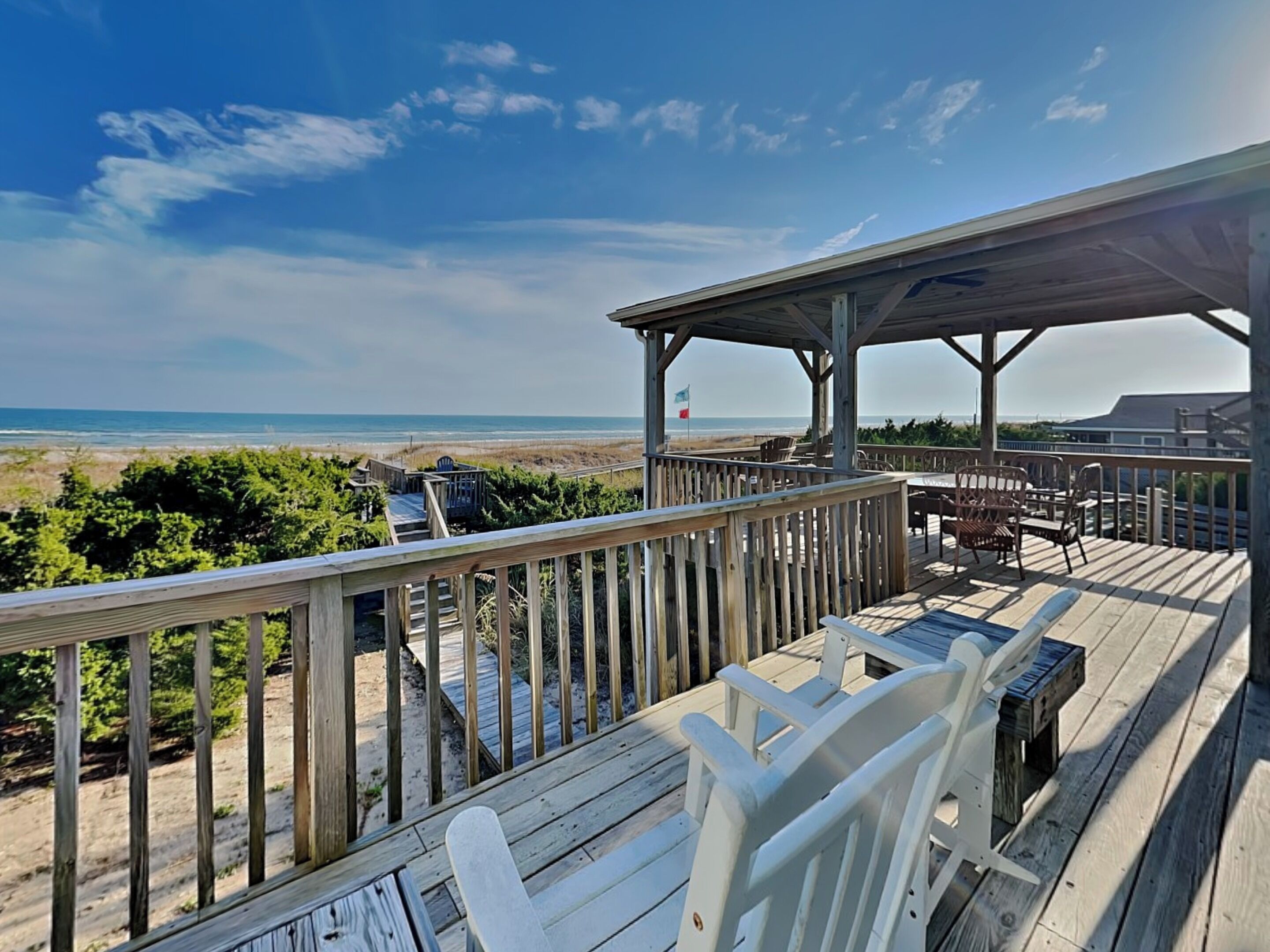 A wooden deck with white chairs and a table overlooking a beach with a flag.