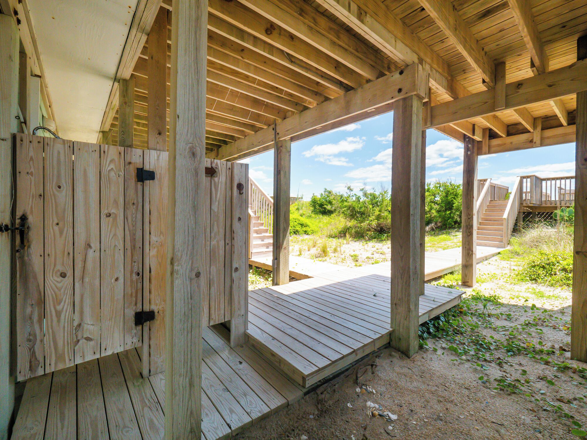 Une structure en bois dotée d’une terrasse couverte et d’un escalier menant à une zone sablonneuse où se trouve de la végétation.