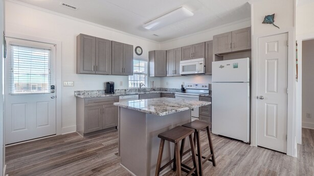 A modern kitchen with grey cabinets, a central island with stools, a microwave, and a refrigerator. - Green Dolphin by Topsail Realty (Holly Ridge)