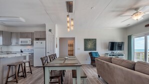 A modern kitchen with a dining area, a living room with a sofa, and a balcony with a TV. - Green Dolphin by Topsail Realty (Holly Ridge)