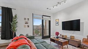 A modern living room with a green sofa, a flat-screen TV, and a large window with a view of buildings. - Mid-Century Charm | Midtown (Nashville)