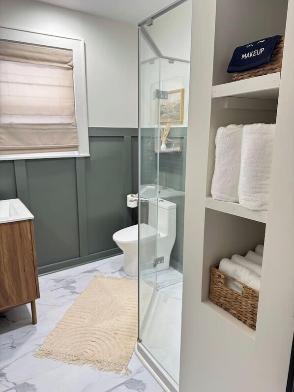 A bathroom with a glass shower enclosure, a toilet, a vanity with a mirror, and a shelf with towels.