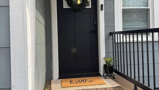 A black front door with a wreath, a doormat, a small potted plant, and a black railing.