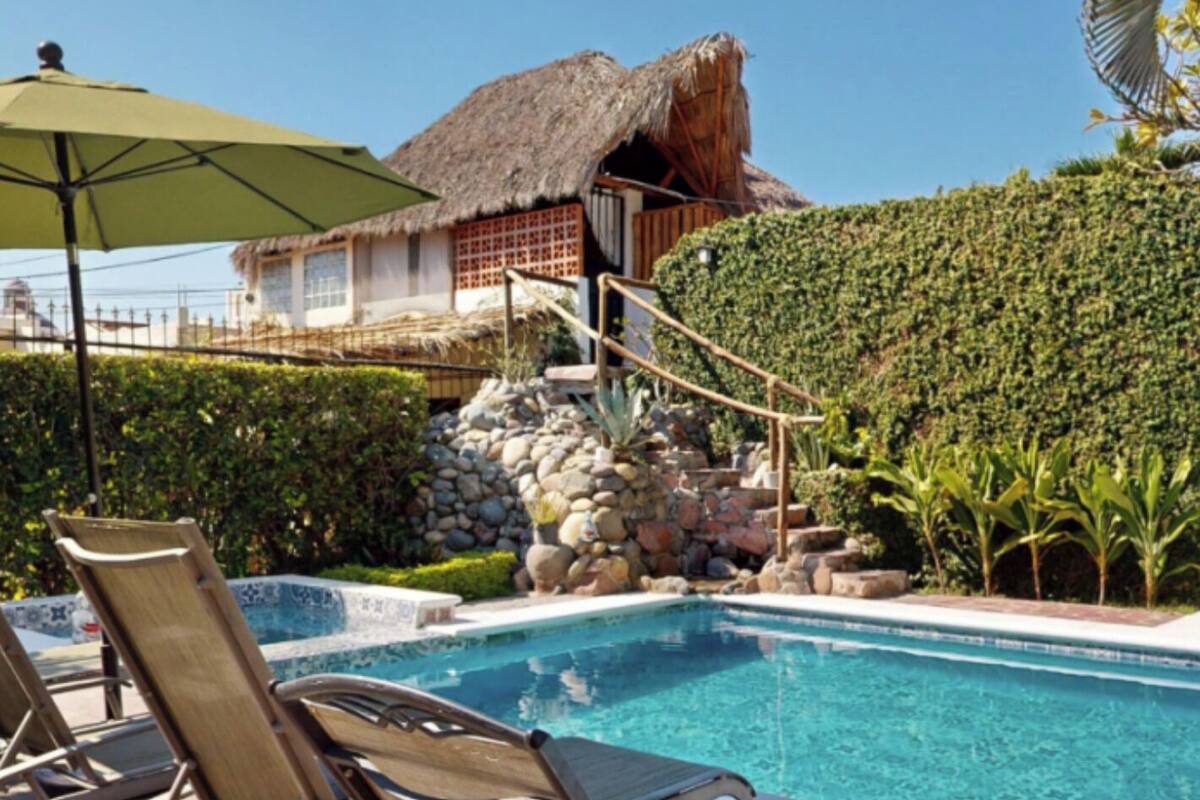 A pool area with lounge chairs, a thatched-roof building, and a stone wall.