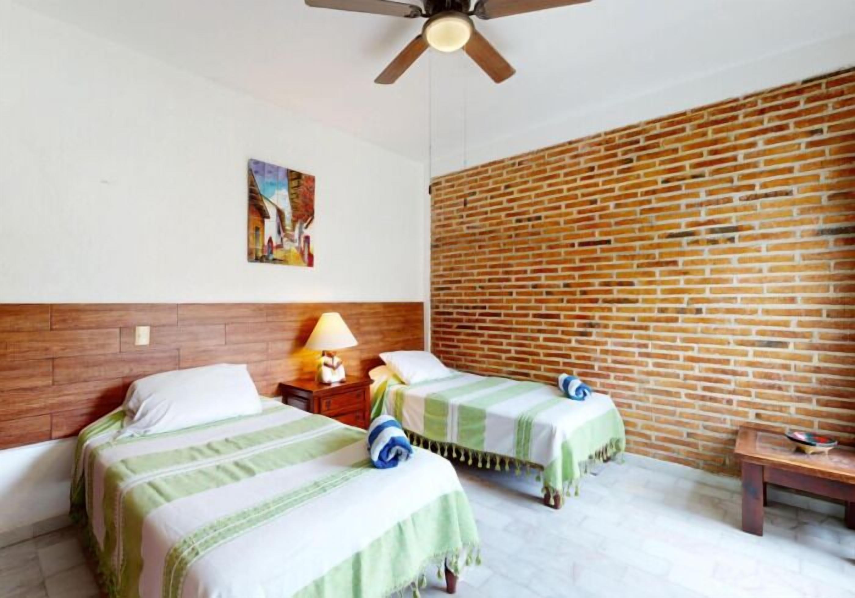 Habitación de hotel con dos camas, una pared de ladrillo, un ventilador de techo y un cuadro en la pared.