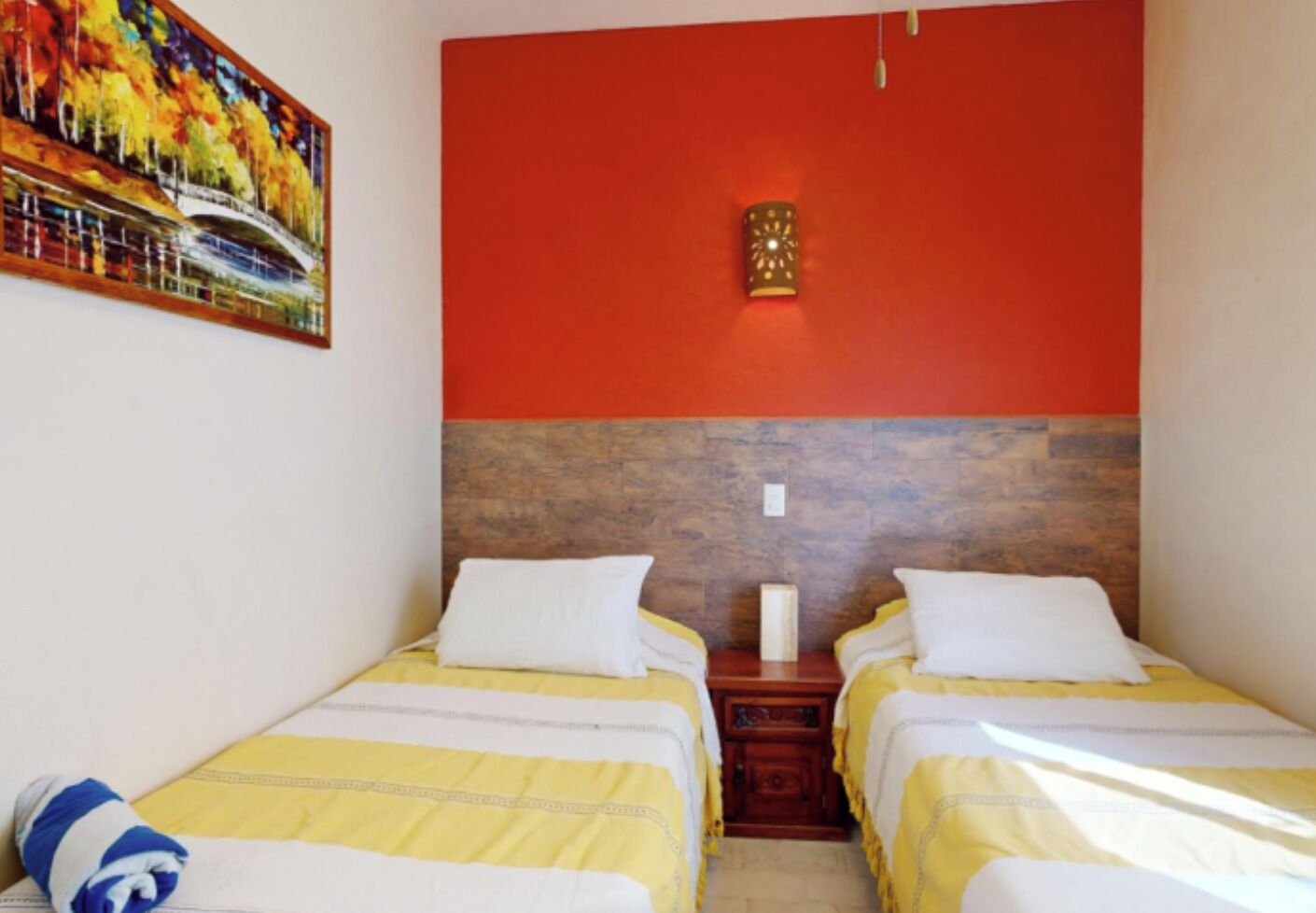 Quarto de hotel com duas camas, cada uma com colcha amarela e branca, cabeceira de madeira e parede de destaque vermelha.