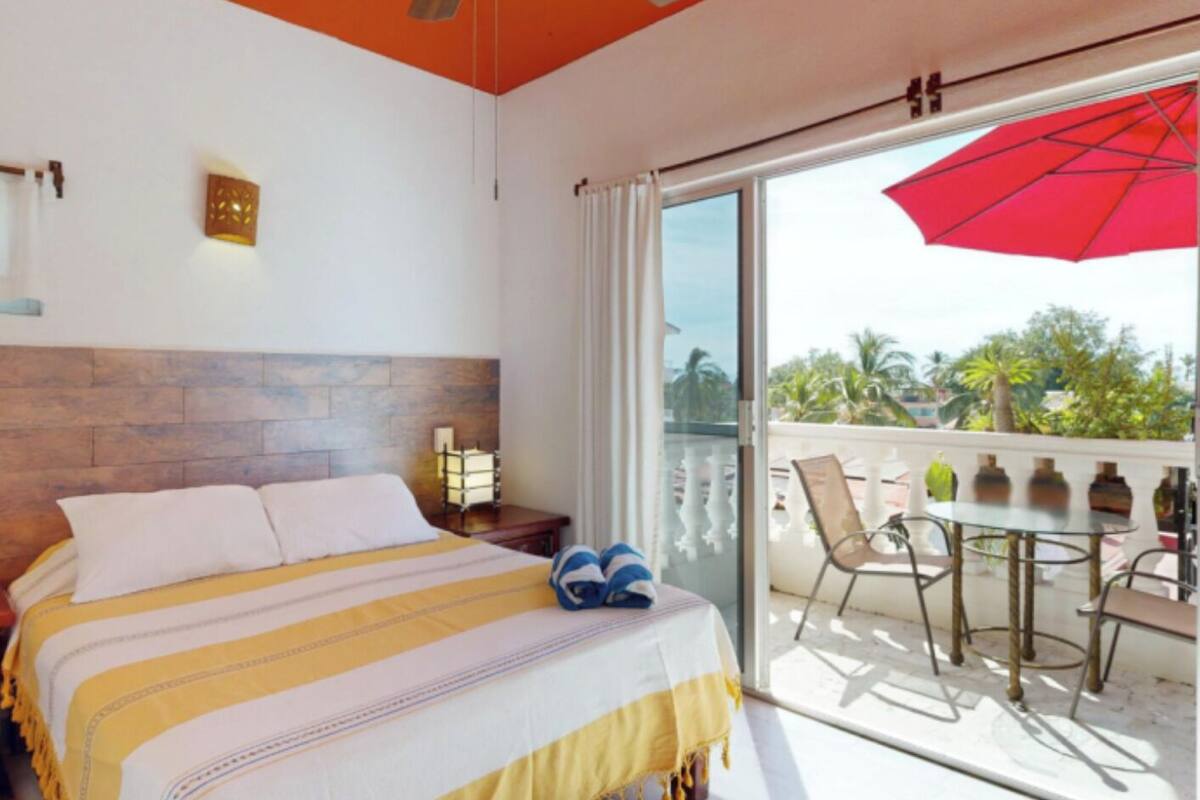 A hotel room with a bed, a nightstand, a lamp, a balcony with a table and chairs, and a red umbrella.