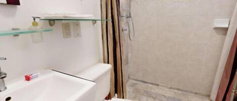 Une salle de bain avec un lavabo blanc, une étagère en verre, une douche avec un rideau et un sol carrelé.