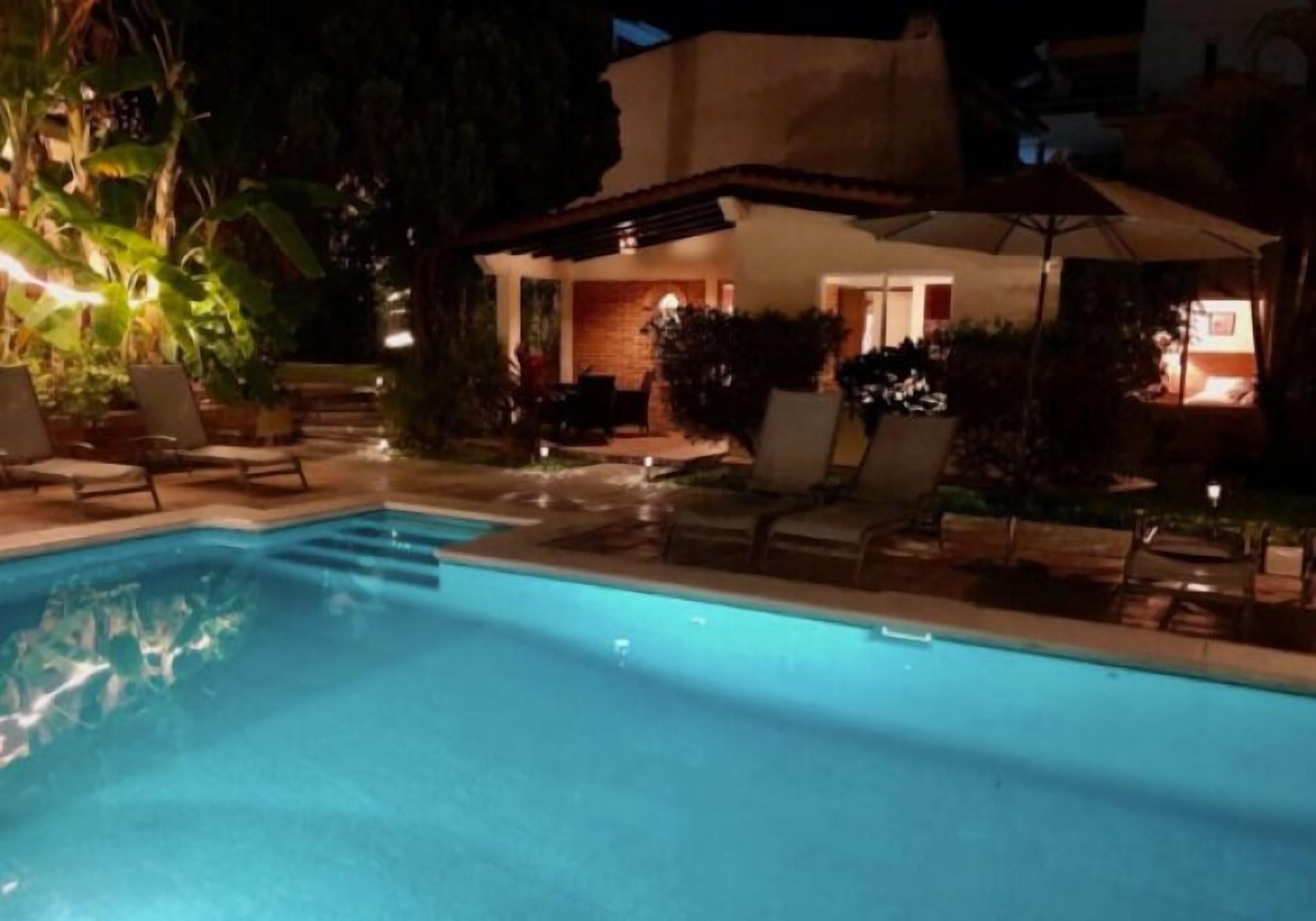 Una piscina con sillas de descanso y sombrillas, rodeada de vegetación y un edificio de fachada de ladrillo.