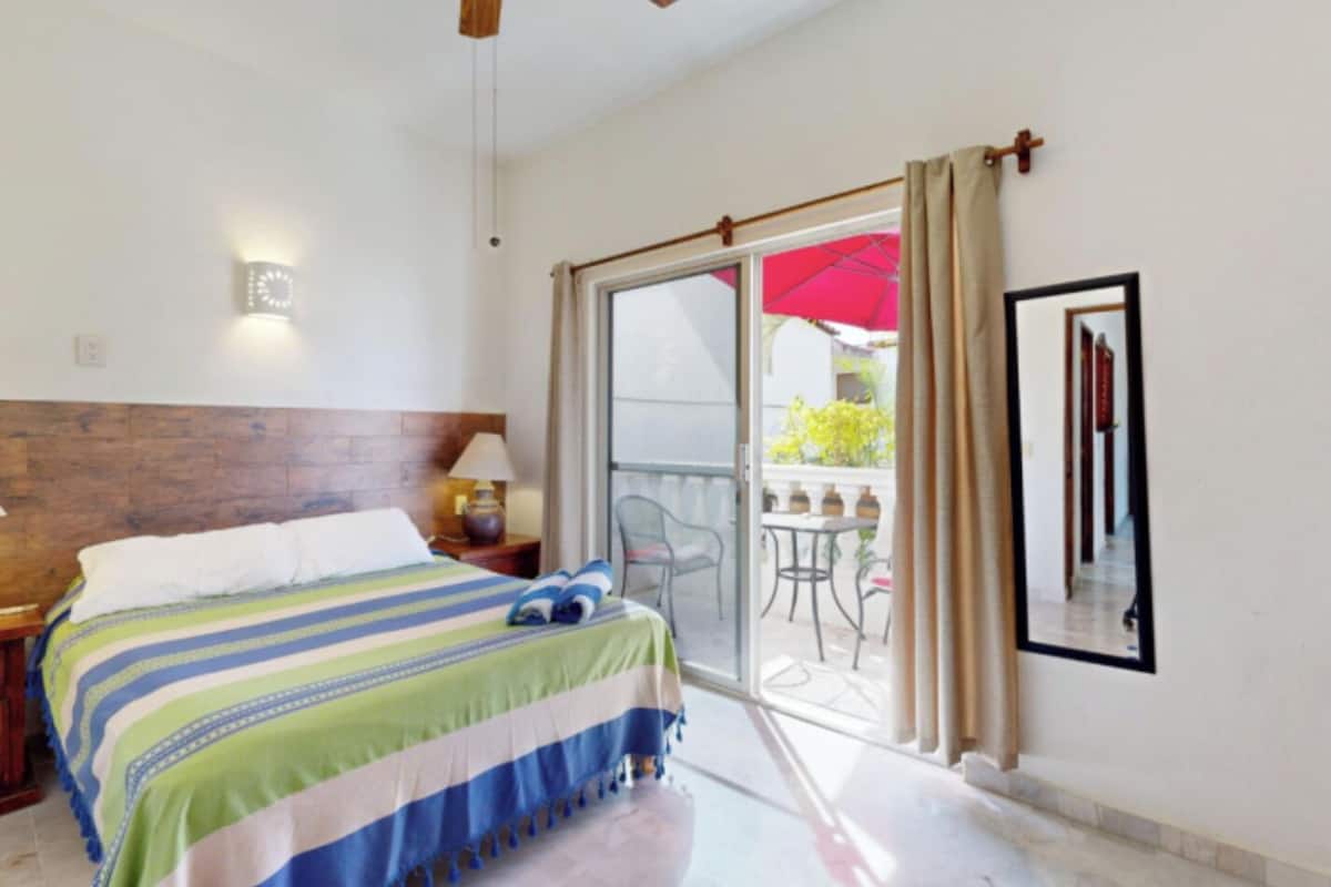 A hotel room with a bed, a nightstand, a lamp, a ceiling fan, and a sliding glass door leading to an outdoor area with a table and chairs.