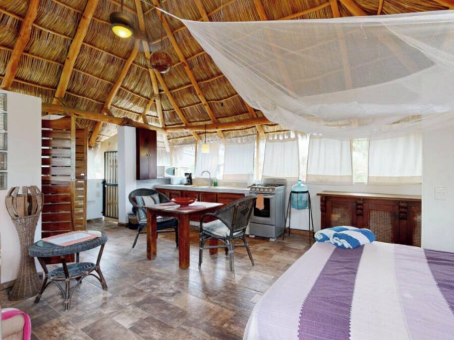 A spacious room with a thatched roof, a dining area with chairs and a table, a bed with a striped blanket, and a kitchenette with a stove and sink.